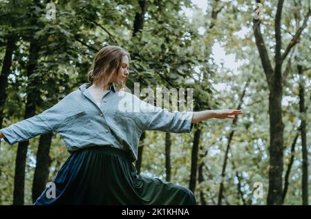 Woman doing yoga warrior II pose in oversized linen shirt with forest background Stock Photo