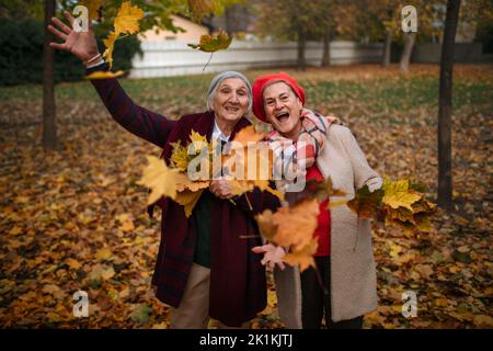 Happy senior female friends on walk outdoors in park in autumn day, having fun and throwing colourful leaves. Stock Photo