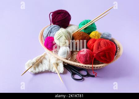 Red, yellow, white, purple, brown, green balls of thread in a beige basket with knitting needles made of bamboo and scissors on a lilac background Stock Photo