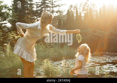 Side view of family standing near pond in park lit up by sunset in summer. Young woman holding grapes over little girl. Stock Photo