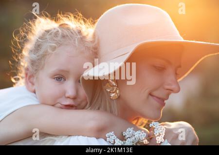 Side view of amazing family standing in forest in summer. Young smiling mother giving piggyback ride to little girl. Stock Photo