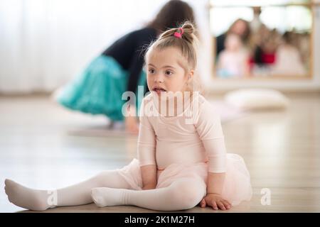 Little girl with down syndrome at ballet class in dance studio,sitting and resting. Concept of integration and education of disabled children. Stock Photo