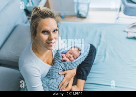 Blond caucasian woman sitting on blue bed in bedroom holding her newborn baby wrapped in blanket looking into camera smiling. Young mother. Horizontal indoor shot. High quality photo Stock Photo