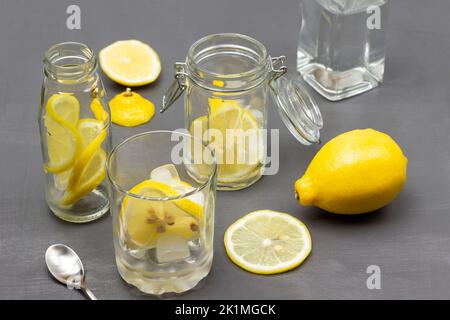 Sliced lemon in glass and in jar. Bottled water. Whole lemon on table. Top view. Grey background. Stock Photo