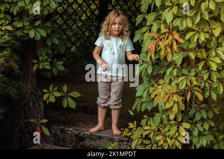 Portrait of cherubic barefoot little girl standing on old stairs in wooden ivy-covered gazebo, playing hide-and-seek. Stock Photo