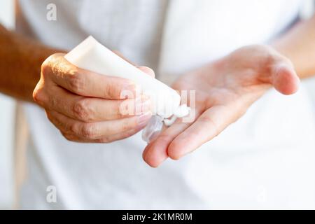 Man skincare. Cosmetic product. Moisturizing treatment. Male hands applying cream from white mockup tube on palm fingers on light blur background. Stock Photo