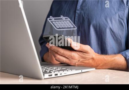 Man using calendar on phone while sitting at table with laptop. Work planning, schedule of events, activities, meetings, agenda preparing. High qualit Stock Photo