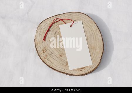 Seasonal sale still life. Blank paper gift tag, label mockup with red rope on cut wooden round board. White linen table cloth background in sunlight Stock Photo