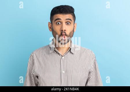 Portrait of funny childish bearded businessman making fish face or sending kiss, having fun, kidding, pretending to be stupid, wearing striped shirt. Indoor studio shot isolated on blue background. Stock Photo