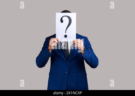 Unknown man hiding his face behind white paper with question mark, finding smart solution, asking for advice, wearing official style suit. Indoor studio shot isolated on gray background. Stock Photo