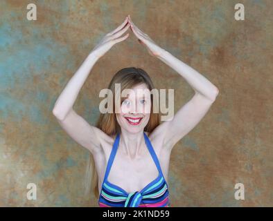 Young woman in striped colorful top smiling and holding her hands in triangle shape over head Stock Photo