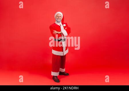 Full length photo of elderly man with gray beard wearing santa claus costume with confident look, standing with folded arms and looking at camera. Indoor studio shot isolated on red background. Stock Photo