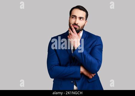 Pensive thoughtful bearded businessman holding his chin with hand, pondering about future, planning strategy, wearing official style suit. Indoor studio shot isolated on gray background. Stock Photo