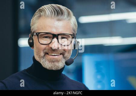 Closeup portrait of senior mature boss with headset, video call, man smiling and looking at camera, gray haired businessman wearing glasses manager investor working inside modern office building, Stock Photo