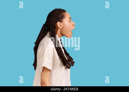 Side view portrait of happy surprised beautiful woman with black dreadlocks notices something incredible, stares amazed, keeps mouth opened. Indoor studio shot isolated on blue background. Stock Photo