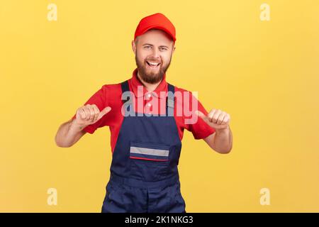 Portrait of happy delighted worker man standing and pointing at himself, being satisfied of result of his work, wearing overalls and red cap. Indoor studio shot isolated on yellow background. Stock Photo