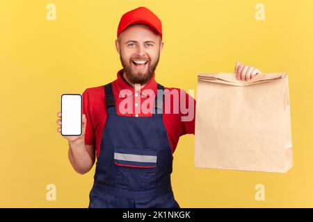 Joyful satisfied deliveryman holding paper parcel and showing smart phone with blank display for advertisement, looking at camera with smile. Indoor studio shot isolated on yellow background. Stock Photo