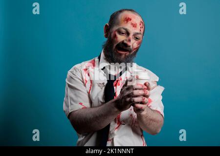 Dangerous zombie looking at cup of coffee, wanting to dirnk beverage and being aggressive in studio. Scary evil monster holding cup and having bloody wounds, apocalyptic eerie devil. Stock Photo