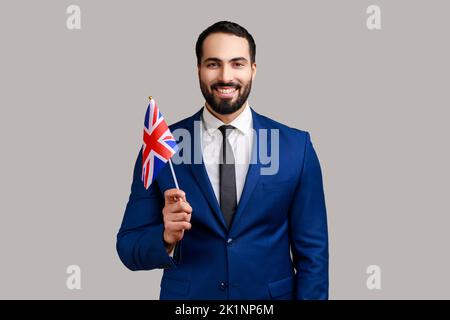 Delighted bearded man holding British flag, celebrating holiday, looking at camera with toothy smile, wearing official style suit. Indoor studio shot isolated on gray background. Stock Photo