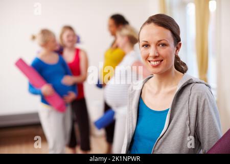 Relaxation exercise in perfect for pregnancy. A young pregnant woman standing in a gym holding an exercise mat with a group of women in the background Stock Photo