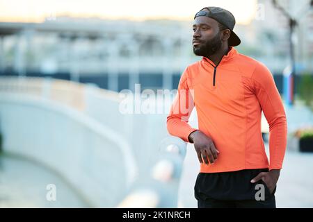 Young serene African American athlete in red sport jacket and black baseball cap standing by riverside and looking at cityscape Stock Photo