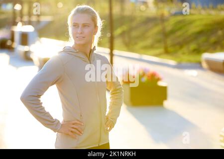 Young serene sportswoman in grey sport jacket keeping hands on waist while standing in front of camera and having break after workout Stock Photo