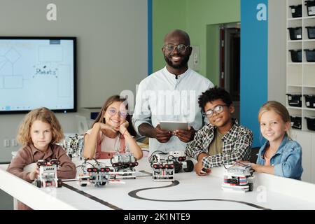 Group of happy youthful intercultural schoolkids and their teacher looking at camera while playing with new models of robots at lesson Stock Photo