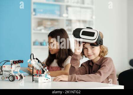 Happy youthful schoolchild taking off vr headset and looking at camera while sitting by desk next to his classmate using tablet Stock Photo