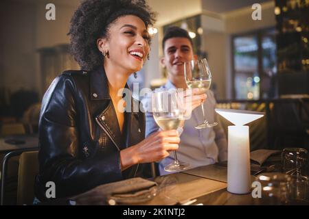 Multiethnic couple of young people sitting at restaurant and having fun talking with friends carefree holding white wine - people, dating and food and Stock Photo