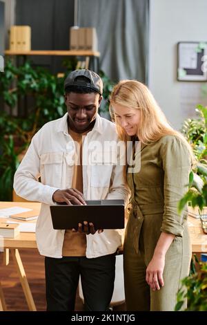 Two young intercultural employees in casualwear looking at laptop screen while standing by desk and preparing presentation of project Stock Photo