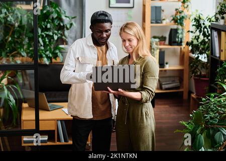 Two young intercultural employees in casualwear looking at screen of laptop held by blond businesswoman during discussion Stock Photo