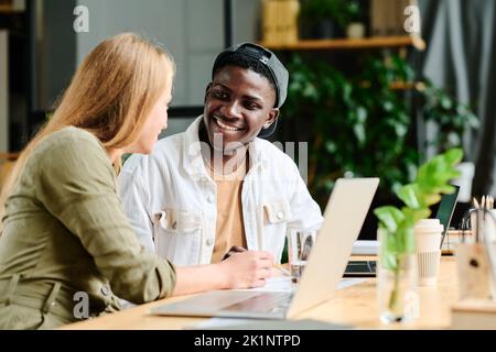 Young smiling African American businessman listening to blond confident female colleague explaining him data which they analyzing Stock Photo
