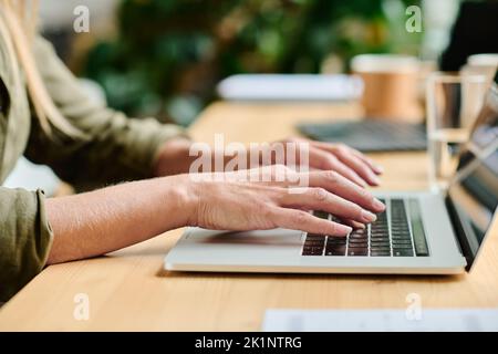 Side view of hands of young businesswoman typing on laptop keyboard while sitting by table in office or cafe and working over project Stock Photo
