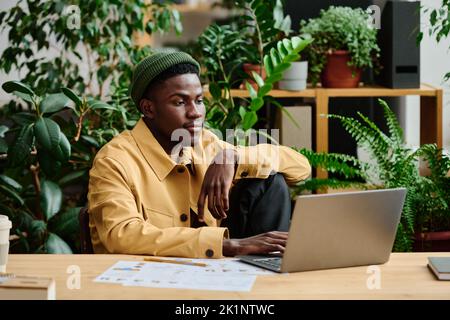 Young serious black man in casualwear sitting by workplace in front of laptop screen in green office and working over business project Stock Photo