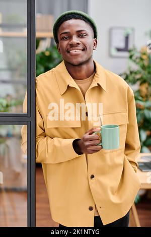 Young successful African American businessman with cup of coffee standing in front of camera against openspace office Stock Photo