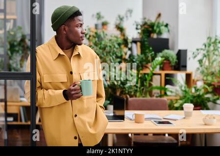 Young black man in stylish casualwear holding cup of coffee while standing in front of camera against his workplace in office Stock Photo