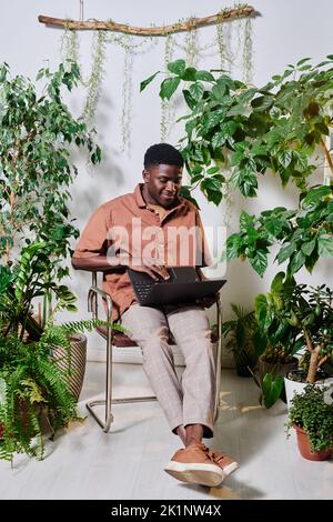 Young African American businessman with laptop on his knees sitting on chair among green plants in modern office and networking Stock Photo