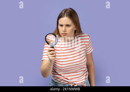 Portrait of blond woman wearing striped T-shirt looking away through magnifying glass, finding out something, exploring, inspecting. Indoor studio shot isolated on purple background. Stock Photo