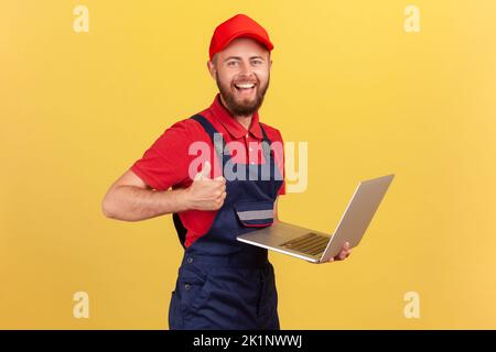 Satisfied worker man wearing blue overalls, red T-shirt and cap working on laptop, showing thumb up, likes a new online service, looking at camera. Indoor studio shot isolated on yellow background. Stock Photo