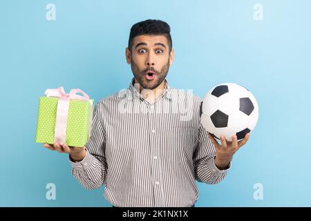 Portrait of shocked surprised businessman standing looking at camera with amazement, holding soccer ball and present box, wearing striped shirt. Indoor studio shot isolated on blue background. Stock Photo