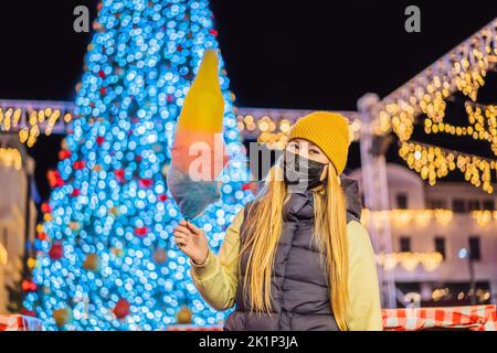 Young woman holding a cotton candy and smiling at a Christmas fair wearing a yellow wool cap wears a medical mask against coronavirus COVID 19 Stock Photo