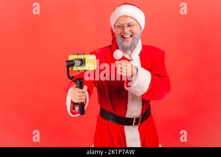 Elderly blogger man with gray beard wearing santa claus costume broadcasting livestream, using phone and steadicam, pointing finger to device display. Indoor studio shot isolated on red background. Stock Photo