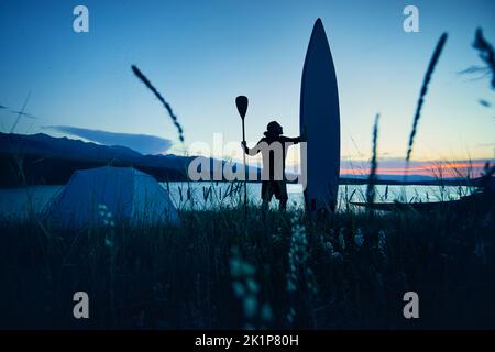 Man in silhouette with paddle and SUP board near tent camping near lake at sunset in Kazakhstan. Stand up paddle boarding outdoor active recreation in Stock Photo