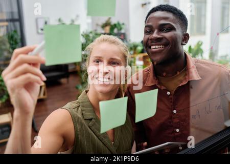 Young smiling blond female employee explaining her colleague working points written on notepapers on transparent noticeboard Stock Photo