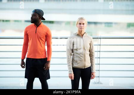 Young multicultural runners or athletes in activewear standing on bridge against riverside while blond sportswoman looking at camera Stock Photo