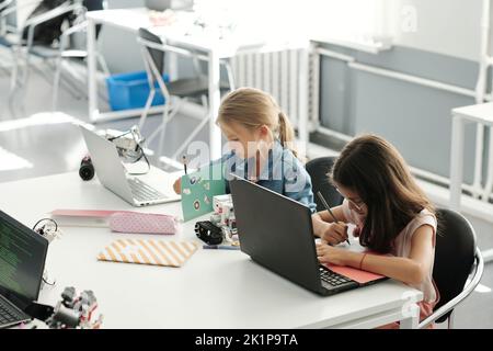 Two clever schoolgirls making notes in their copybooks while sitting in front of laptops by desk and preparing school project Stock Photo