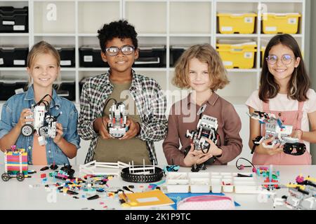 Group of happy youthful intercultural elementary schoolkids holding self made robots constructed at lesson of robotics in school workshop Stock Photo