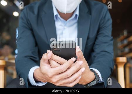 young Businessman in suit wearing surgical face mask and using smartphone, man typing touchscreen mobile phone in office or cafe. Covid-19 Pandemic, t Stock Photo