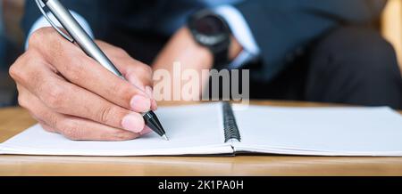 Businessman in suit writing something on notebook in office or cafe, hand of man holding pen with signature on paper report. business concepts Stock Photo
