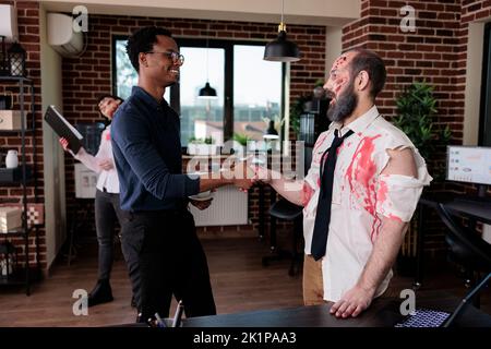 Businessman doing handshake with evil zombie, having conversation with brain eating corpse in startup office. Undead scary monster with bloody wounds saluting person, sinister devil. Stock Photo